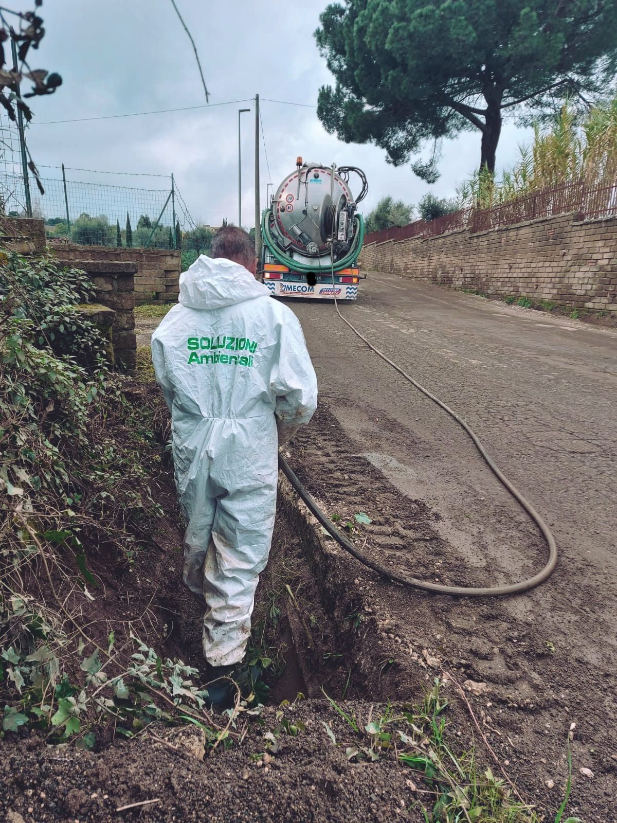 Una persona con una tuta protettiva bianca e un tubo vicino a un camion della nettezza urbana su una strada sterrata.