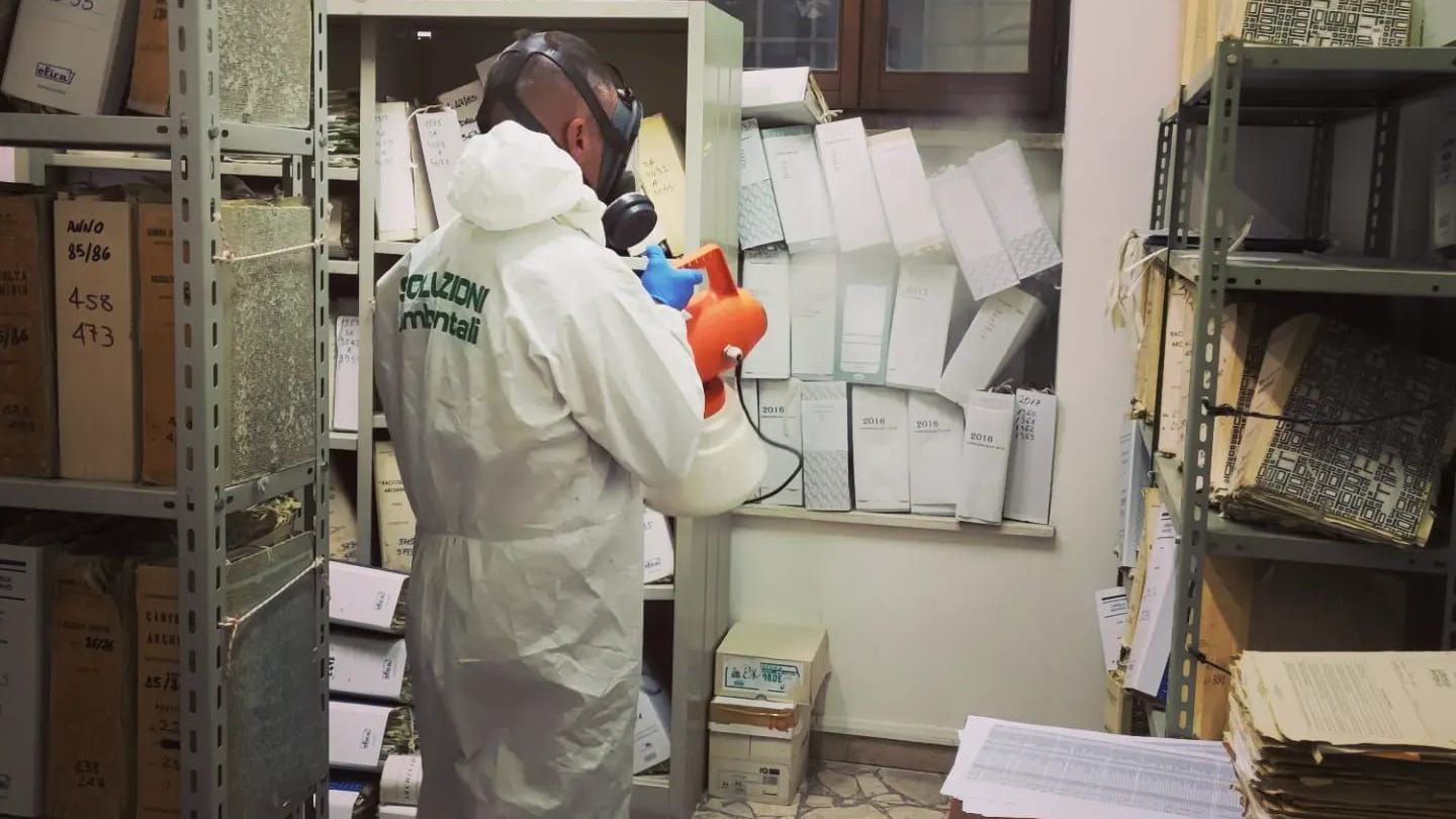 Persona con tuta protettiva che spruzza disinfettante in un archivio pieno di libri e documenti.