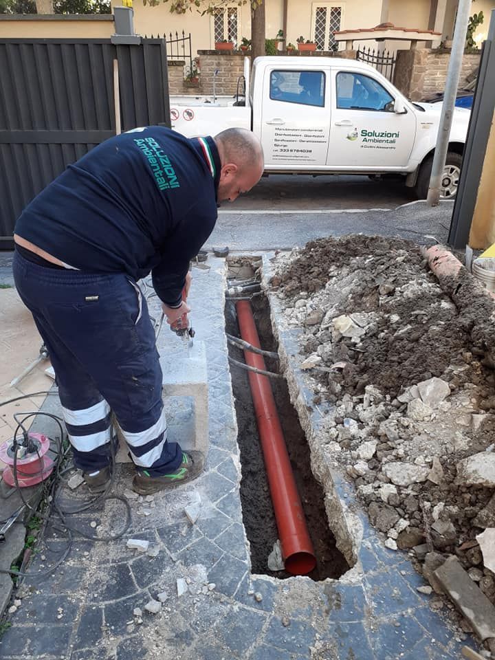 Uomo al lavoro su un tubo arancione interrato in una trincea su un'area asfaltata, con un camion sullo sfondo.