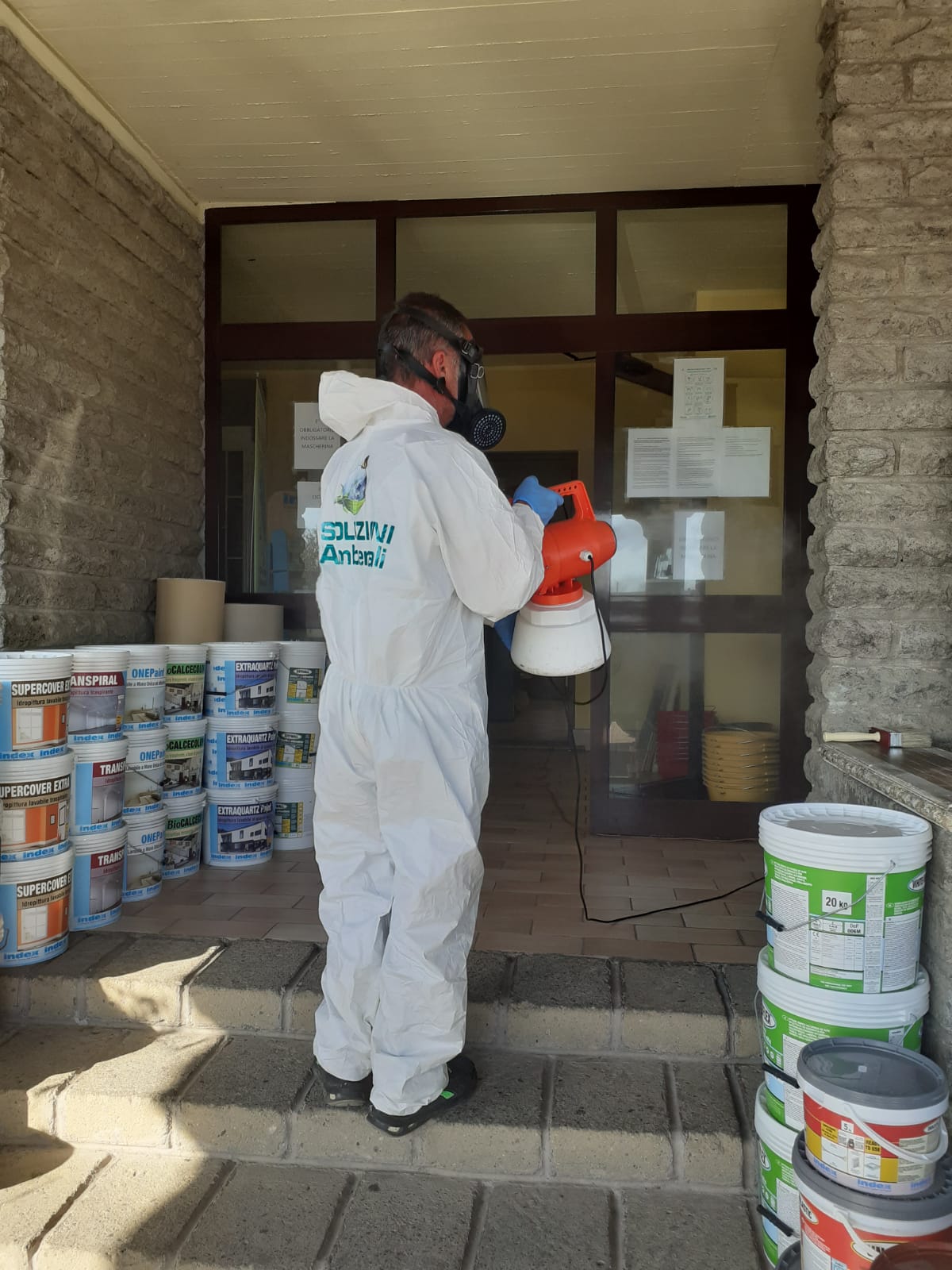 Persona con tuta protettiva che spruzza disinfettante su una porta, circondata da barattoli di vernice.