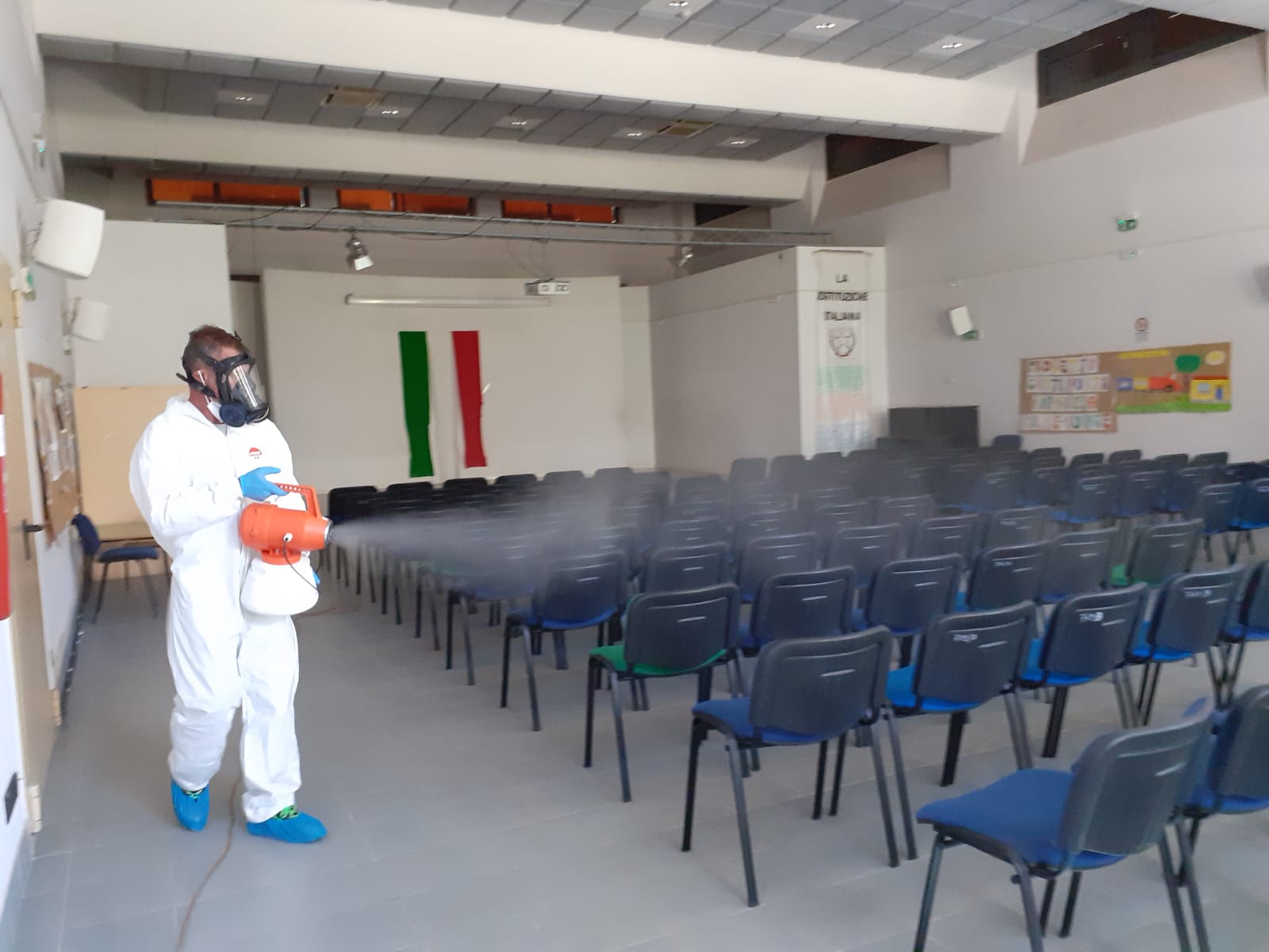 Persona in tuta protettiva che disinfetta sedie in una stanza con una bandiera italiana.