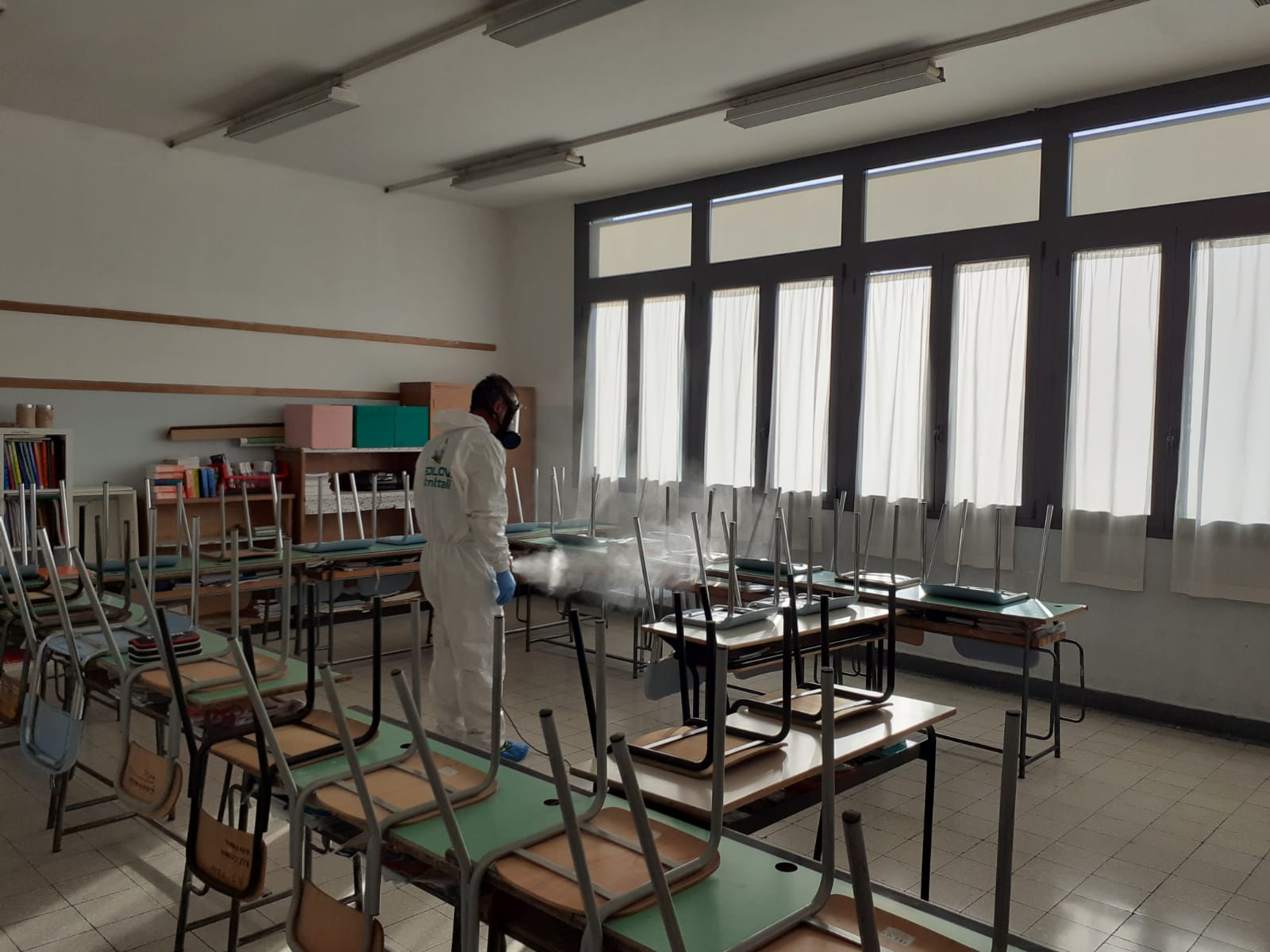 Persona con tuta protettiva che disinfetta un'aula con sedie e banchi rovesciati.