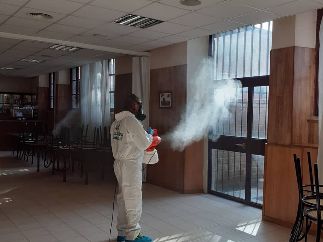 Persona con tuta protettiva che spruzza disinfettante in una stanza con sedie, finestre e tavoli.