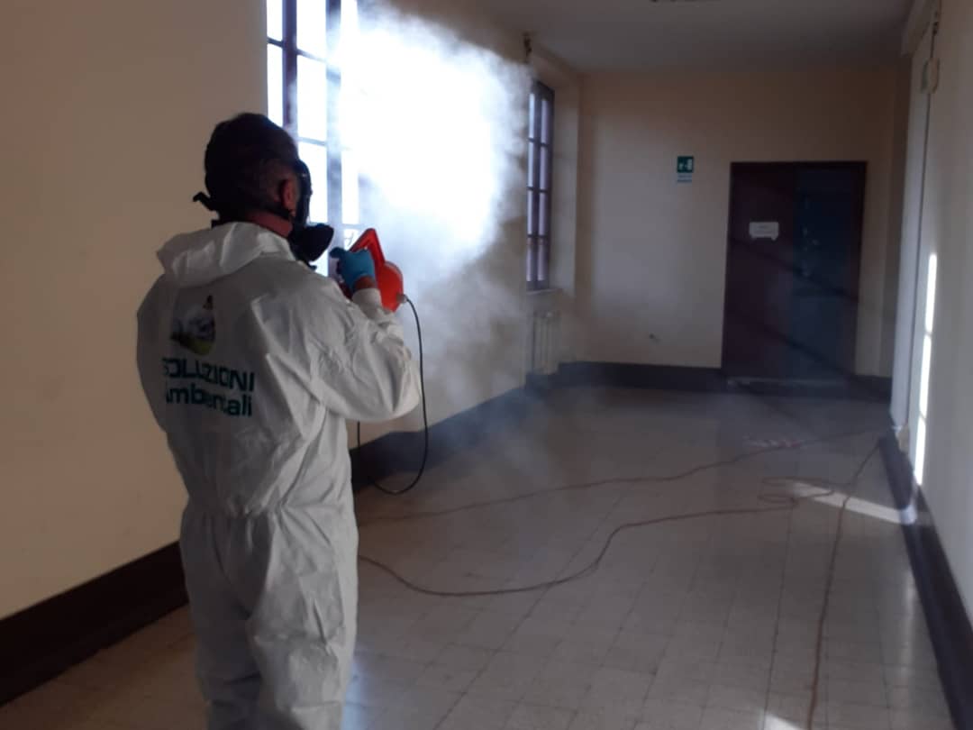 Persona in tuta protettiva che disinfetta un corridoio con un nebulizzatore, spruzzando disinfettante.