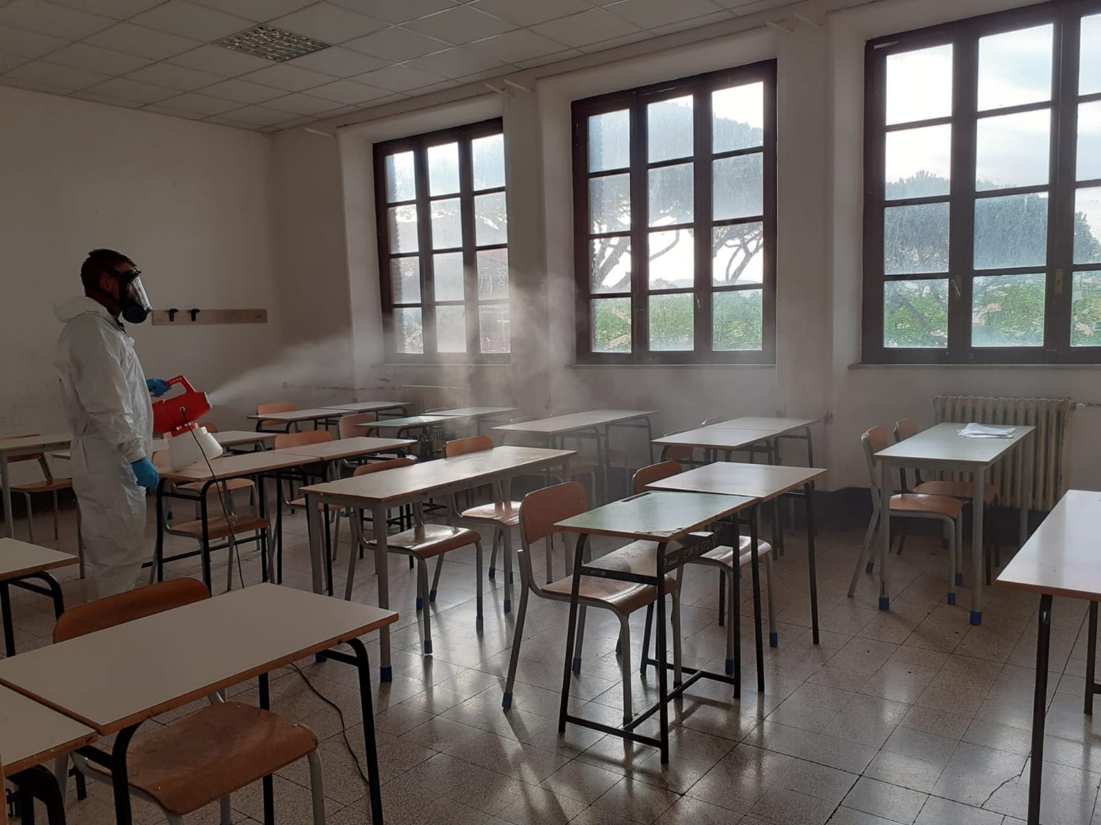 Persona con tuta protettiva che disinfetta un'aula scolastica con banchi e finestre.