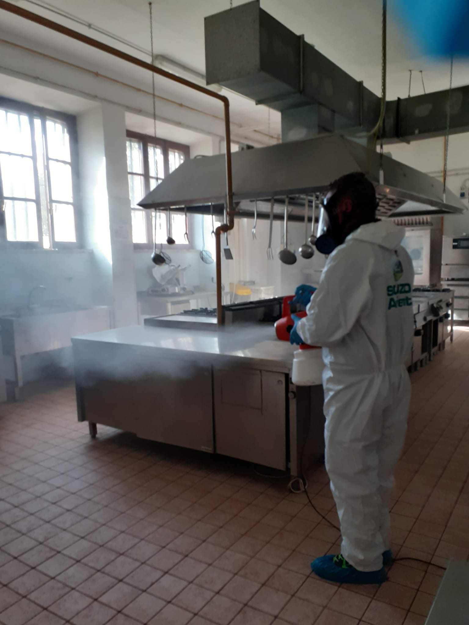 Persona in tuta protettiva che disinfetta una cucina commerciale con un nebulizzatore.