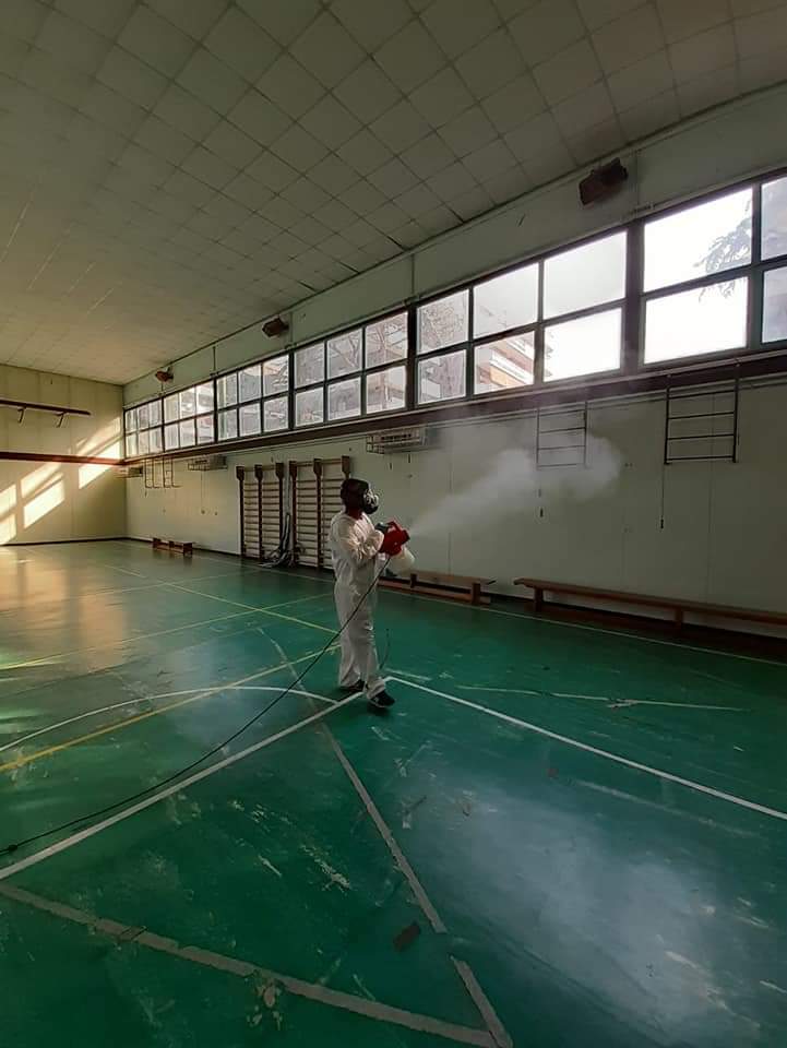Persona in tuta protettiva che disinfetta una palestra con pavimento verde e finestre lungo la parete, utilizzando un nebulizzatore.
