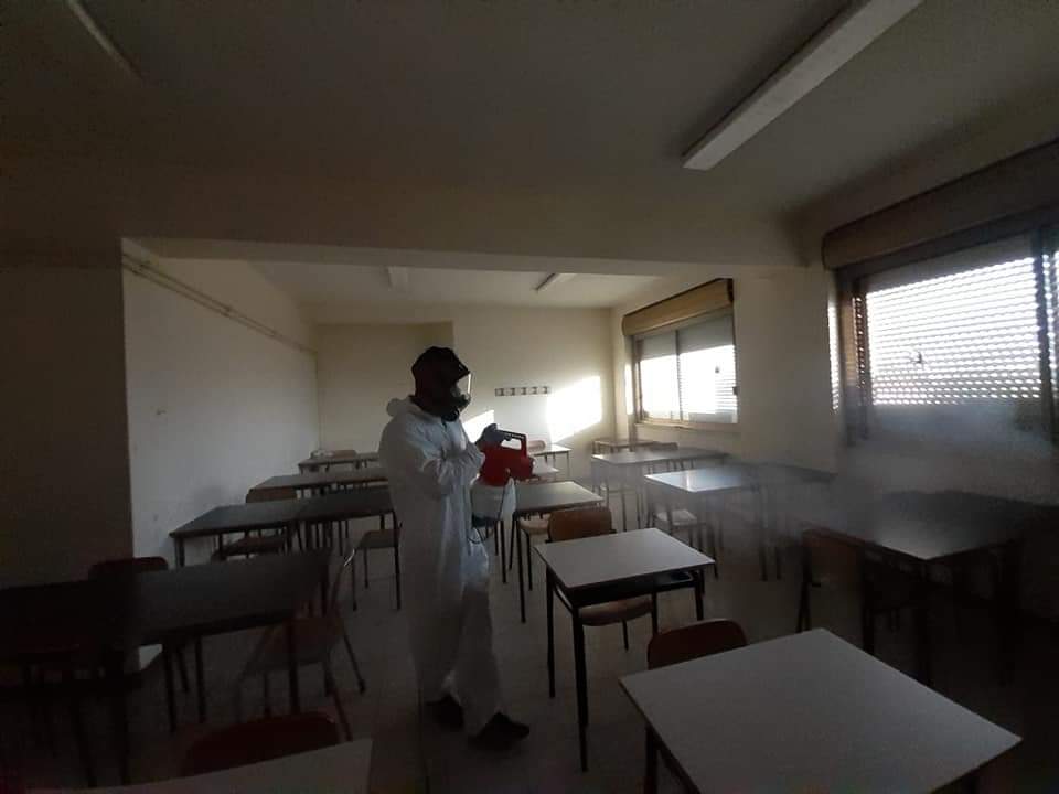 Persona con tuta protettiva che disinfetta un'aula con uno spray.