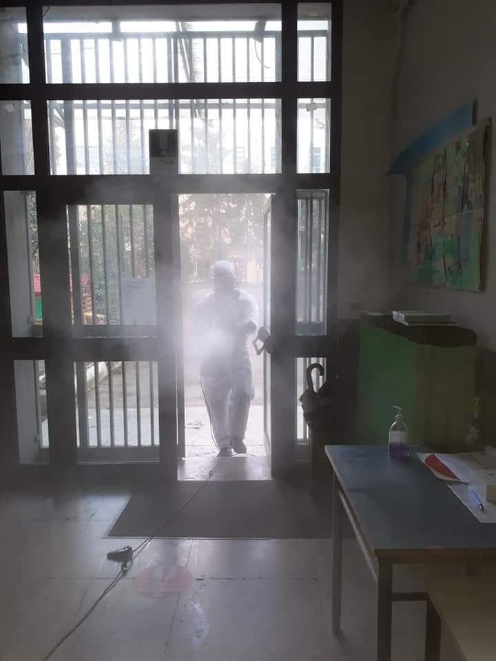 Persona in tuta protettiva che disinfetta una porta con un nebulizzatore. All'interno, con un tavolo e una porta visibili.