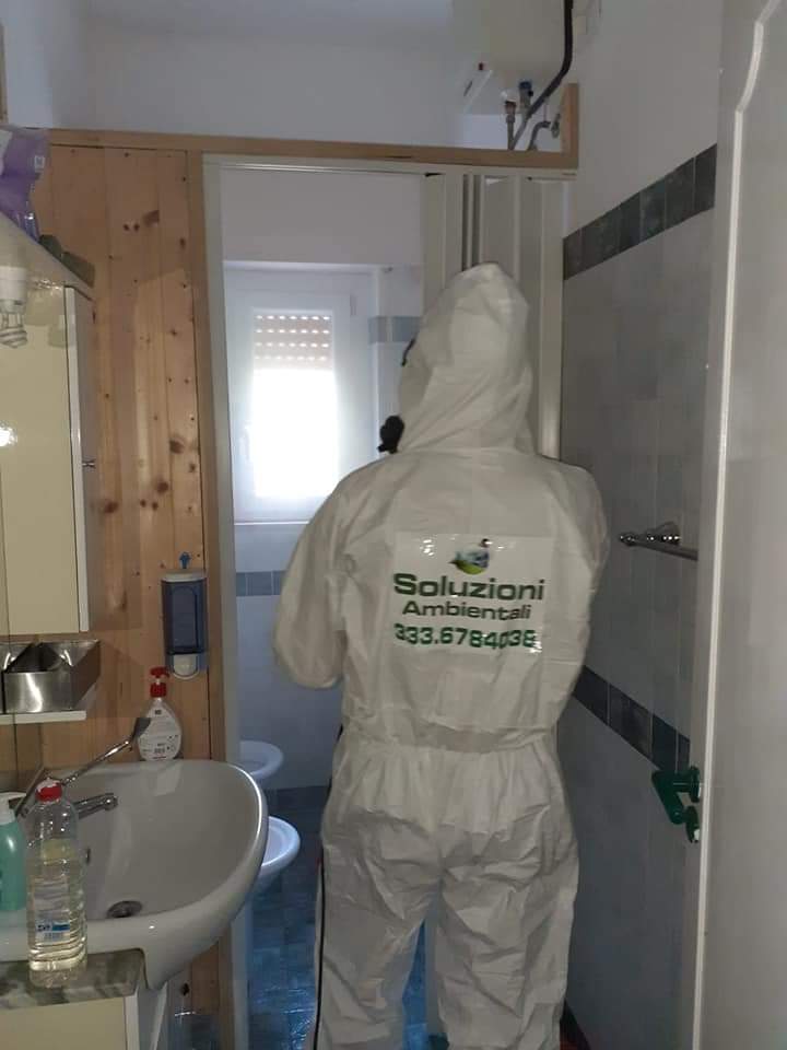 Una persona con una tuta protettiva bianca spruzza acqua in un bagno, rendendo visibili lo stipite della porta e il lavandino.