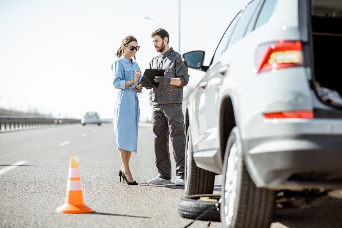 Woman in blue dress and mechanic by a car with a flat tire on the side of the road.