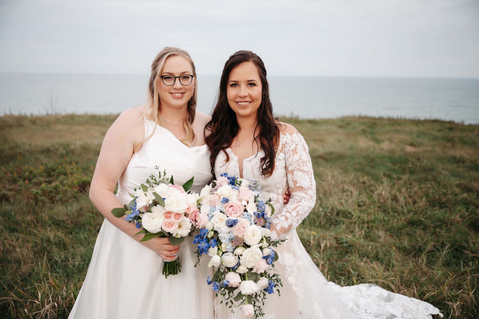 Two women in wedding dresses are standing next to each other in a field holding bouquets of flowers.
