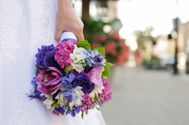 A bride in a white dress is holding a bouquet of purple and pink flowers.