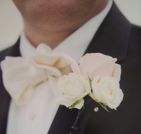 A man in a tuxedo and bow tie has two white roses pinned to his lapel