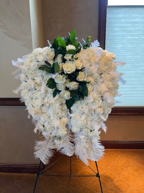 A wreath made of white flowers and feathers is sitting on a stand in front of a window.