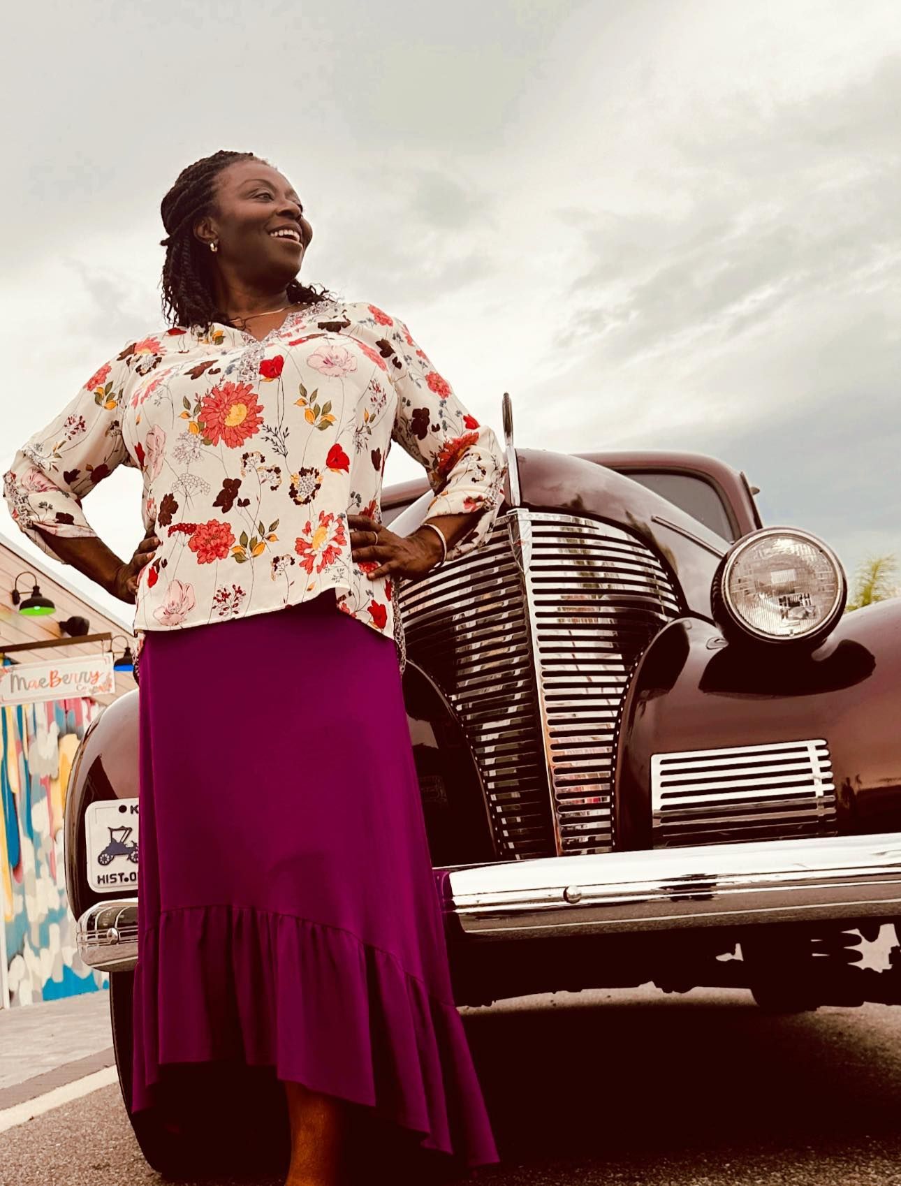 A woman in a purple skirt is standing in front of an old car.