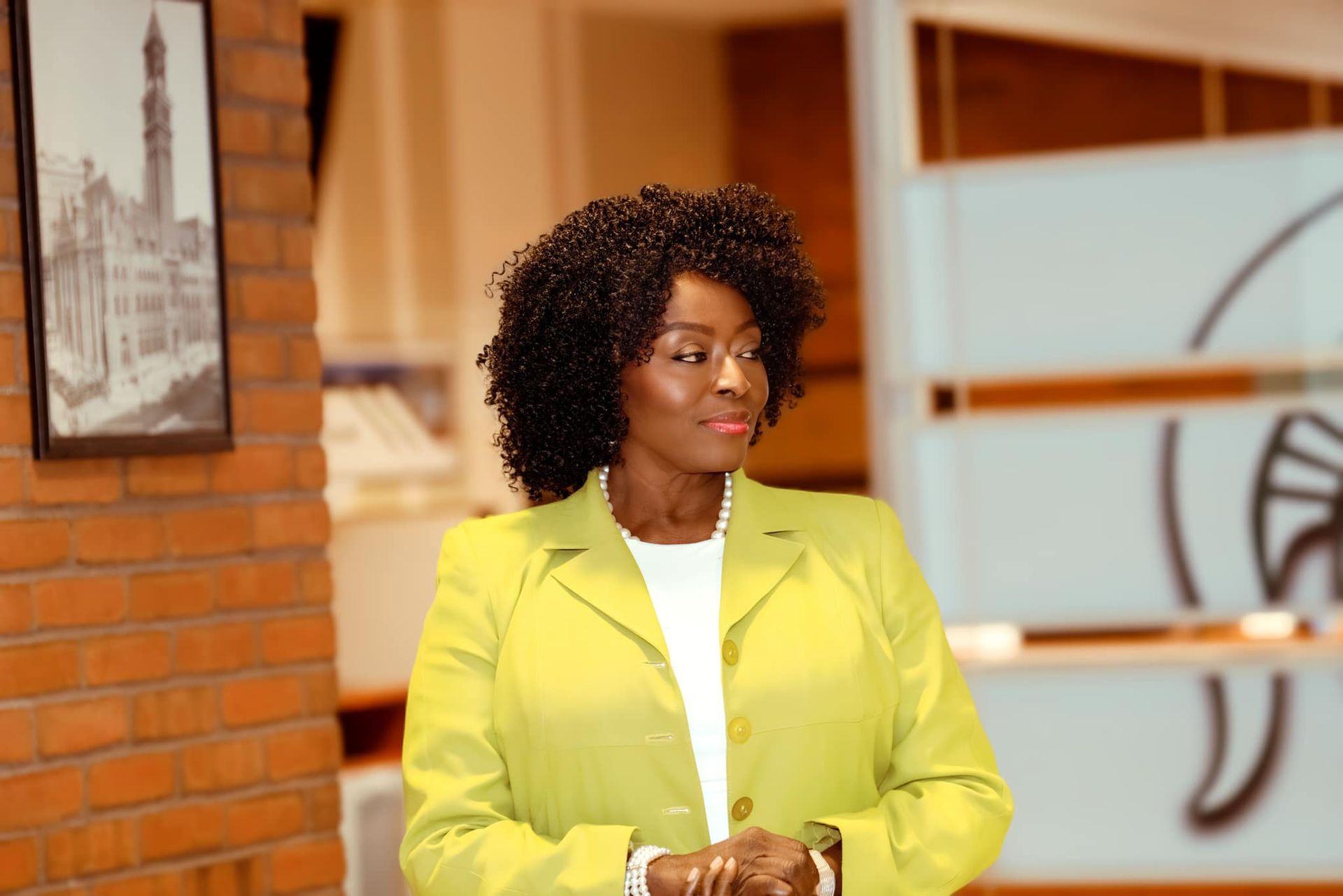 A woman in a yellow jacket is standing in front of a brick wall.