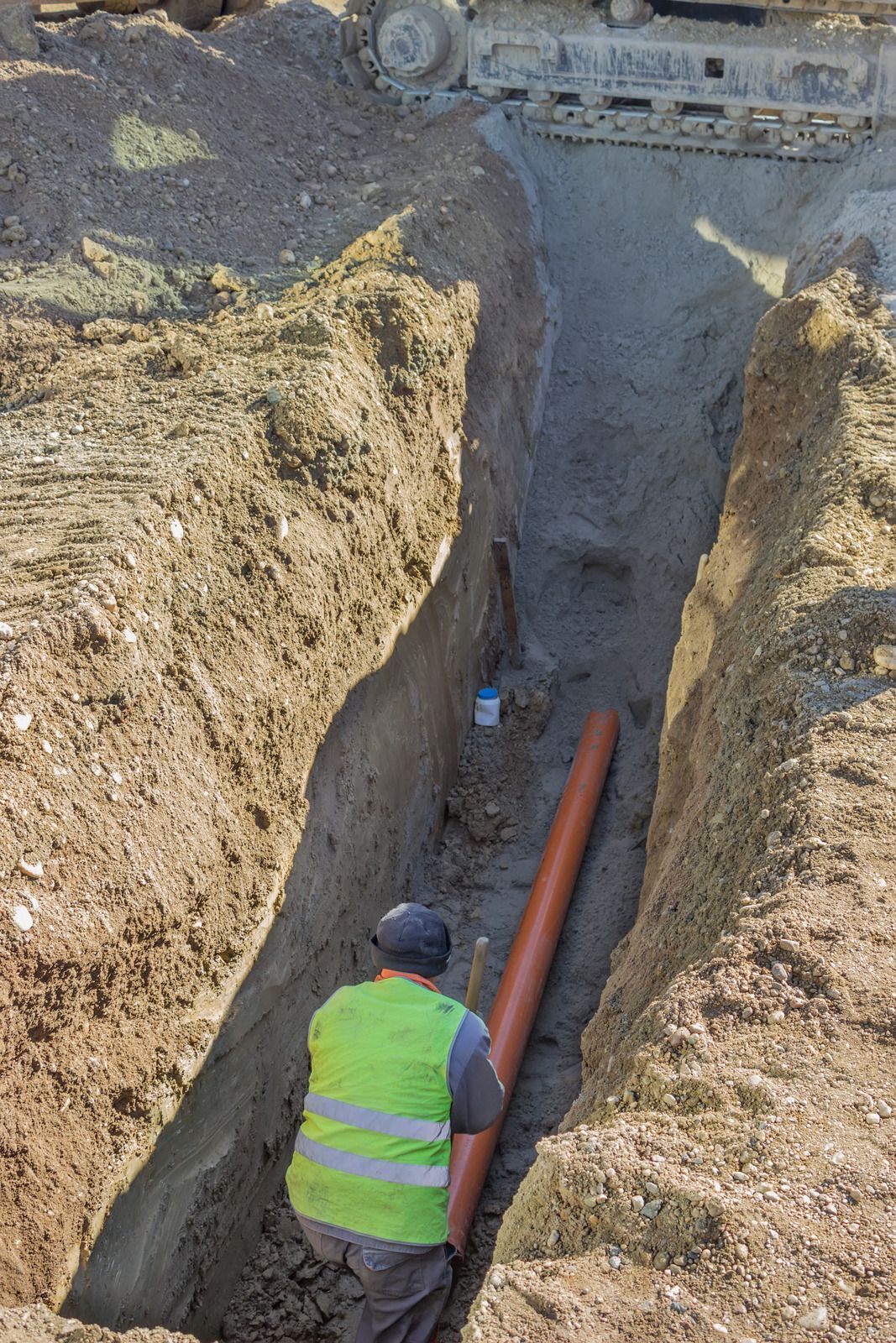 A man in a yellow vest is kneeling in a trench holding a pipe.