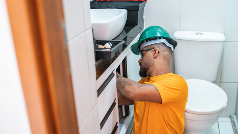 Man in hard hat installing cabinet in bathroom near a toilet.
