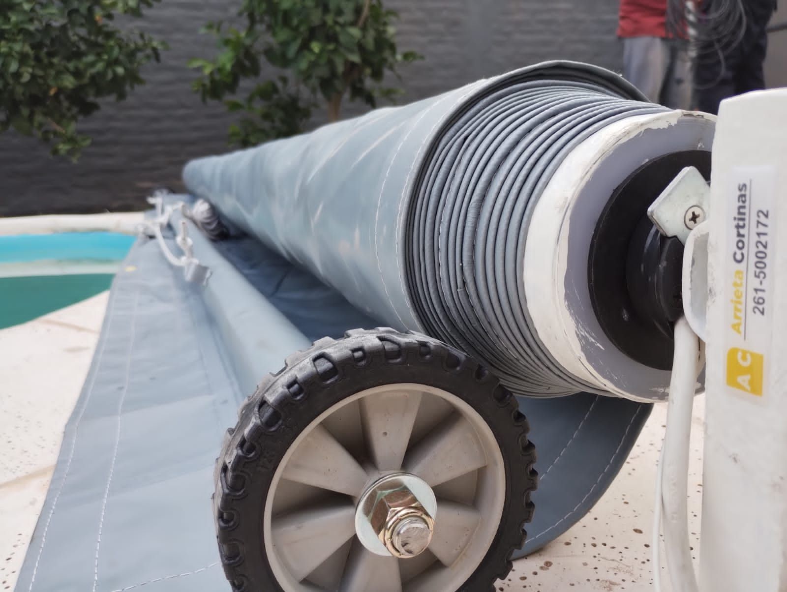 Cubierta de piscina enrollada, tela gris, ruedas y una caja de control blanca cerca de la piscina.