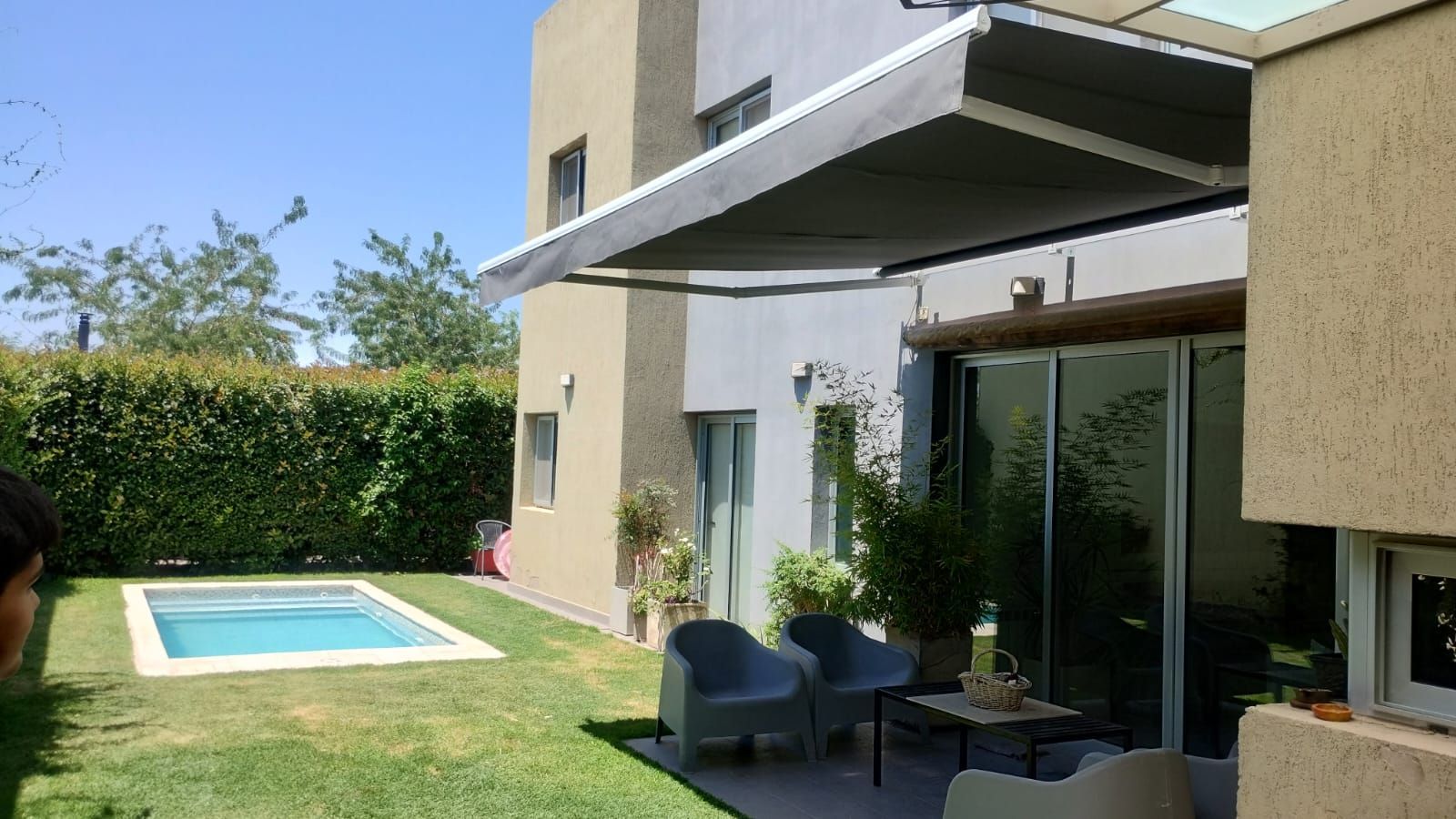 Un toldo retráctil da sombra a un patio con una pequeña piscina y sillas en el césped. Día soleado.