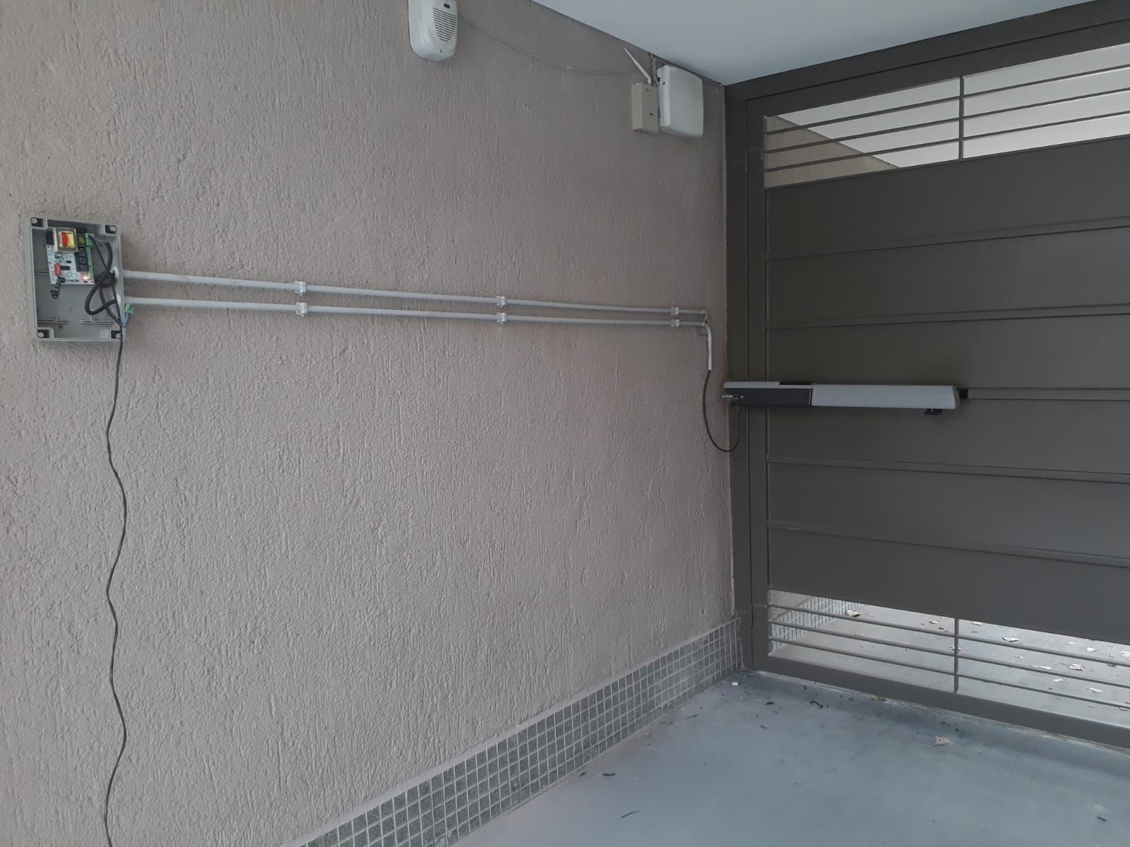 Un sistema de apertura de portón con una caja de control sobre una pared texturizada. Un portón gris está abierto.