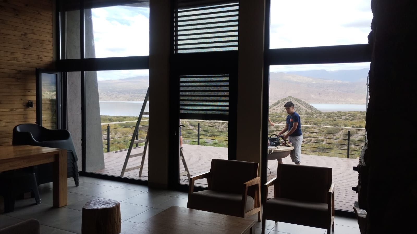 Vista interior de una casa con grandes ventanales con vistas a un paisaje de montaña; una persona está de pie en la terraza.