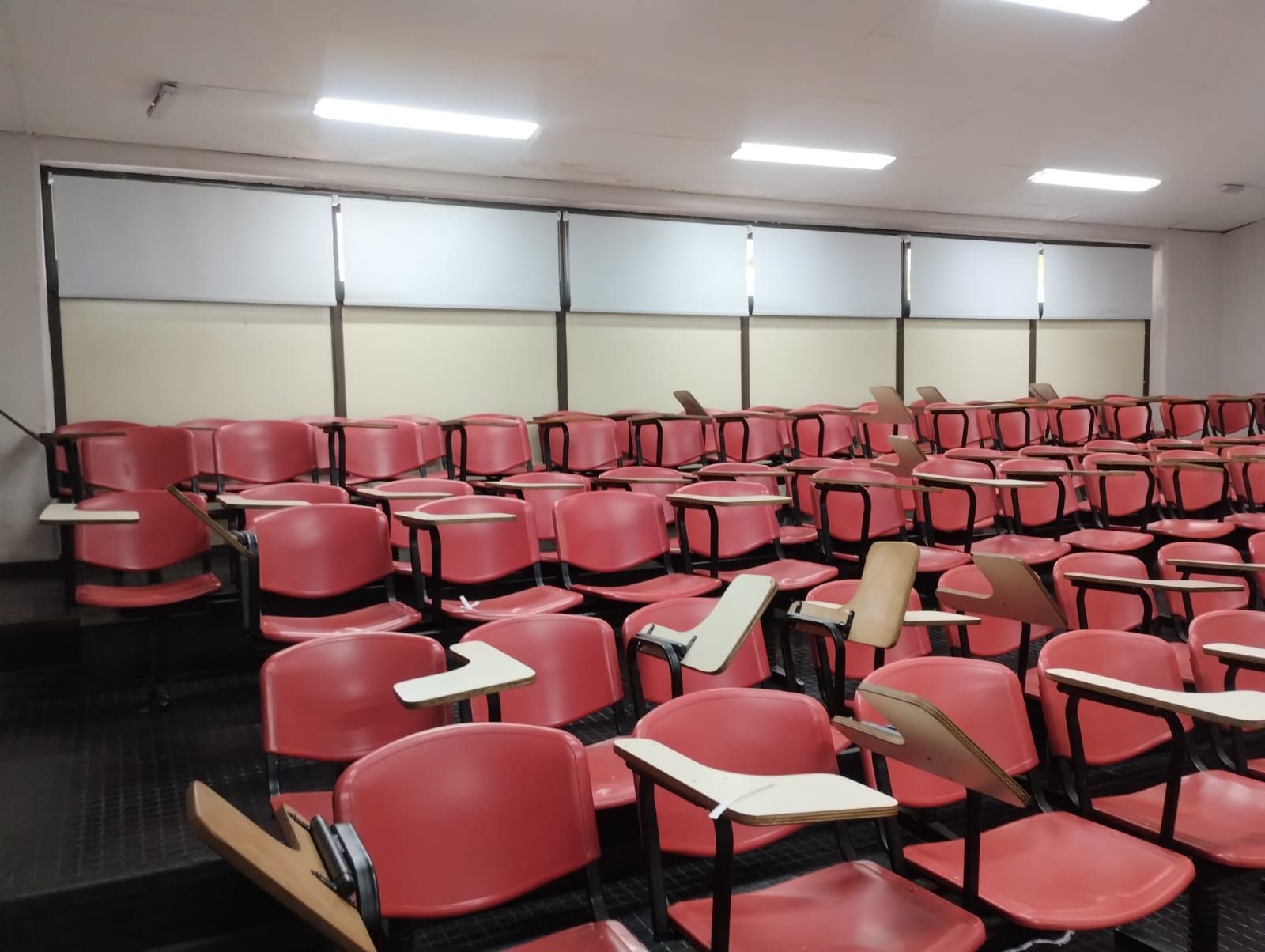Sala de conferencias roja vacía con filas de sillas de escritorio y persianas cerradas.