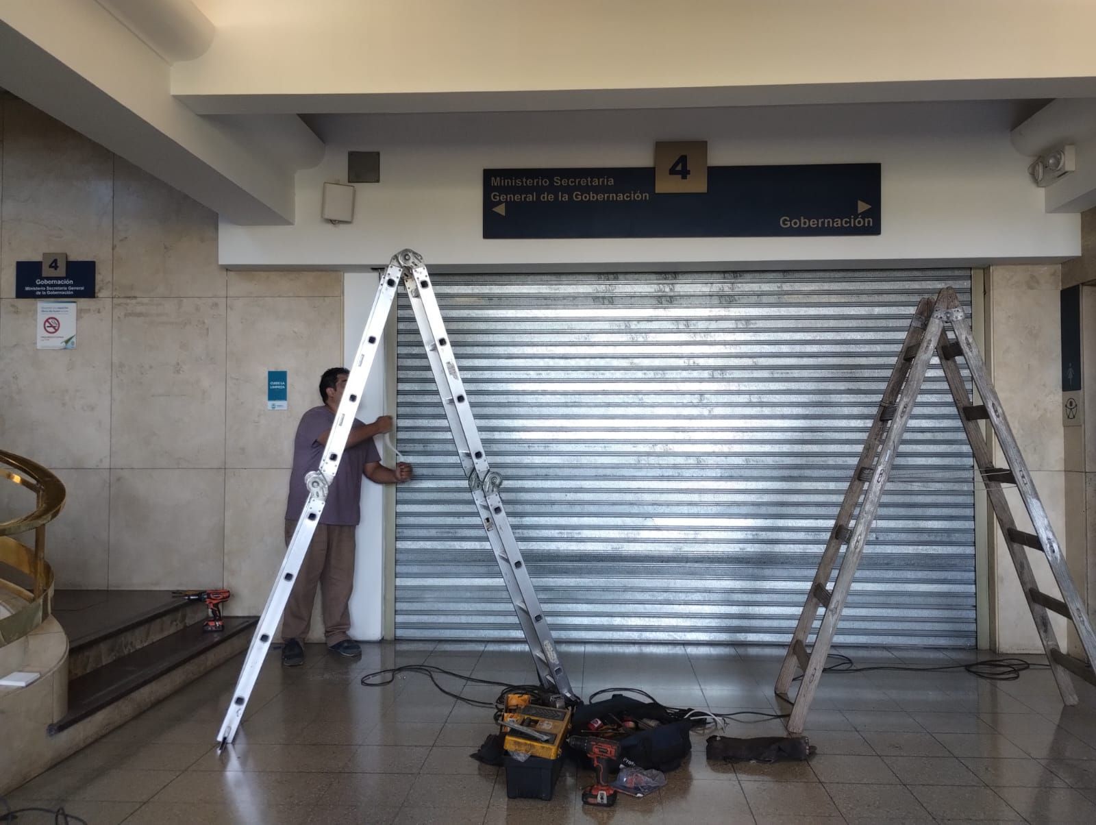 Hombre ajustando una persiana metálica cerrada con dos escaleras apoyadas. Interior.
