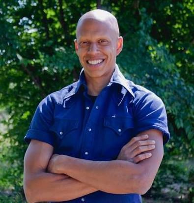 A bald man in a blue shirt is smiling with his arms crossed.