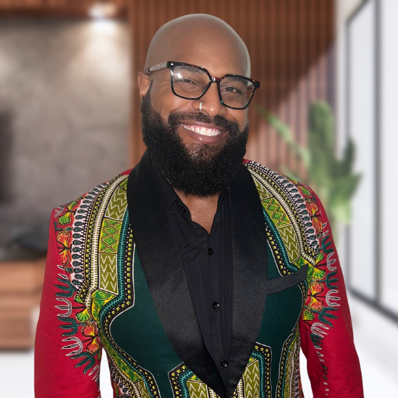 A bald man with a beard is wearing glasses and a colorful jacket.