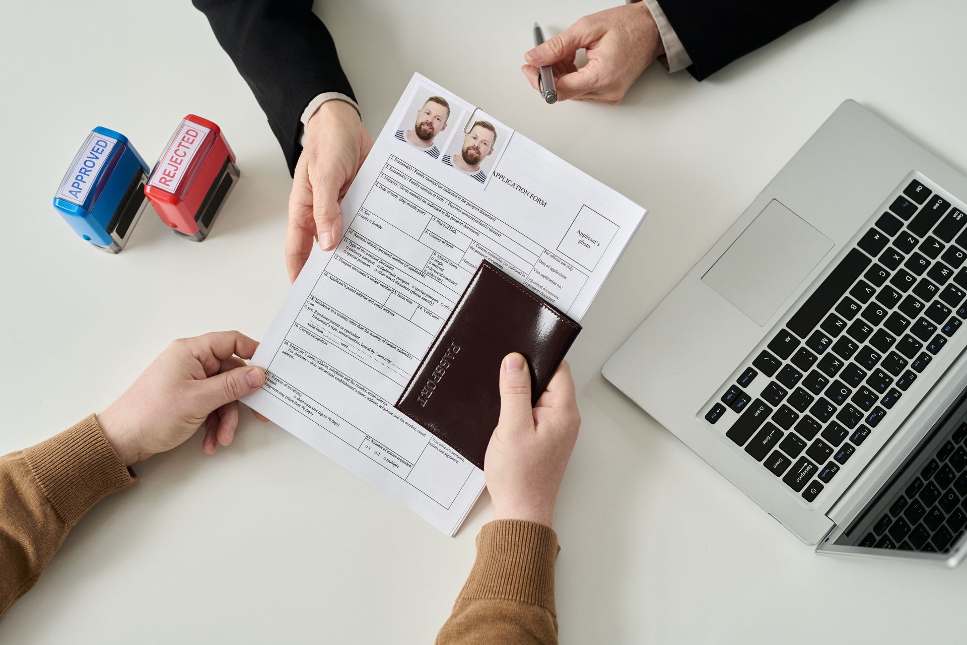 Hands holding passport and document; person behind the desk, laptop and stamps present.