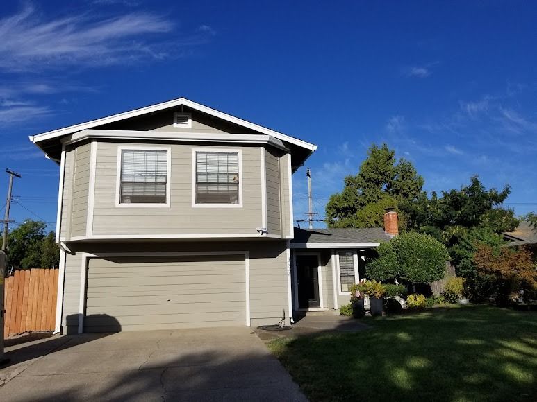 Two-story house with tan siding, gray garage door, green lawn, and blue sky.