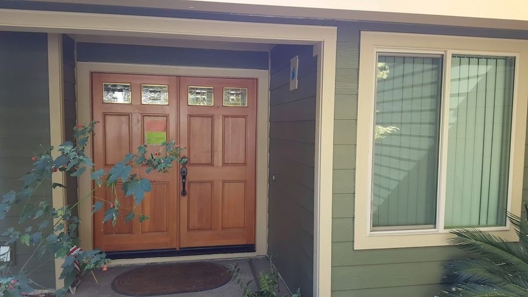 Wooden double doors with a wreath, next to a green-sided house and a window with vertical blinds.