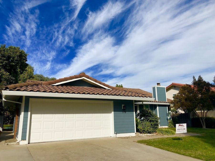 House with blue siding, white garage door, and brown tile roof under a blue sky with wispy clouds.