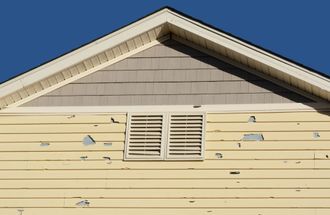 Yellow house siding with peeling paint, a vent, and a gable under a blue sky.