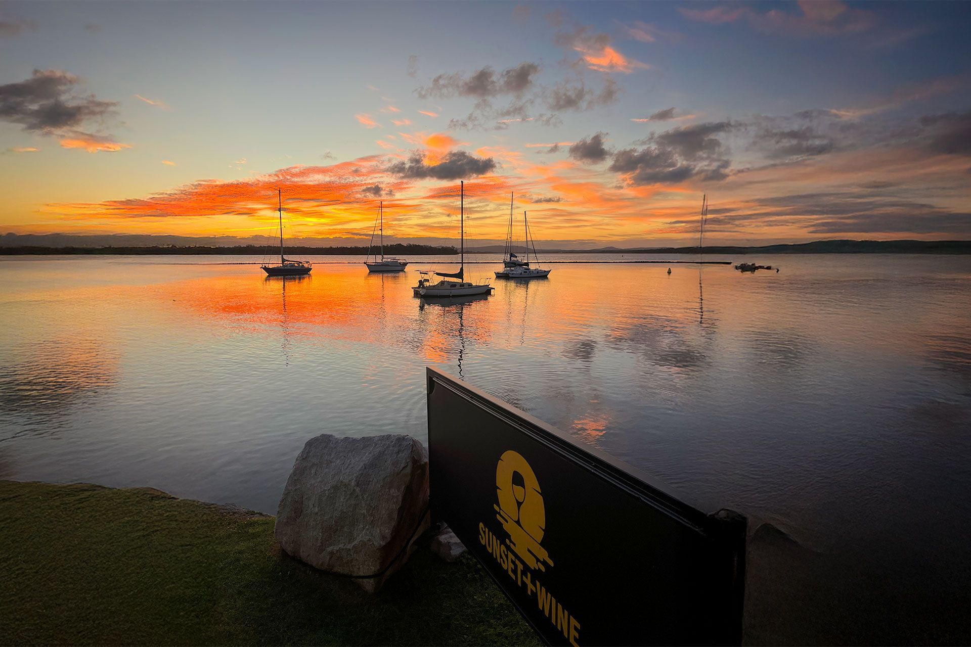Sunset over calm water with sailboats; orange and yellow sky reflected in the water.