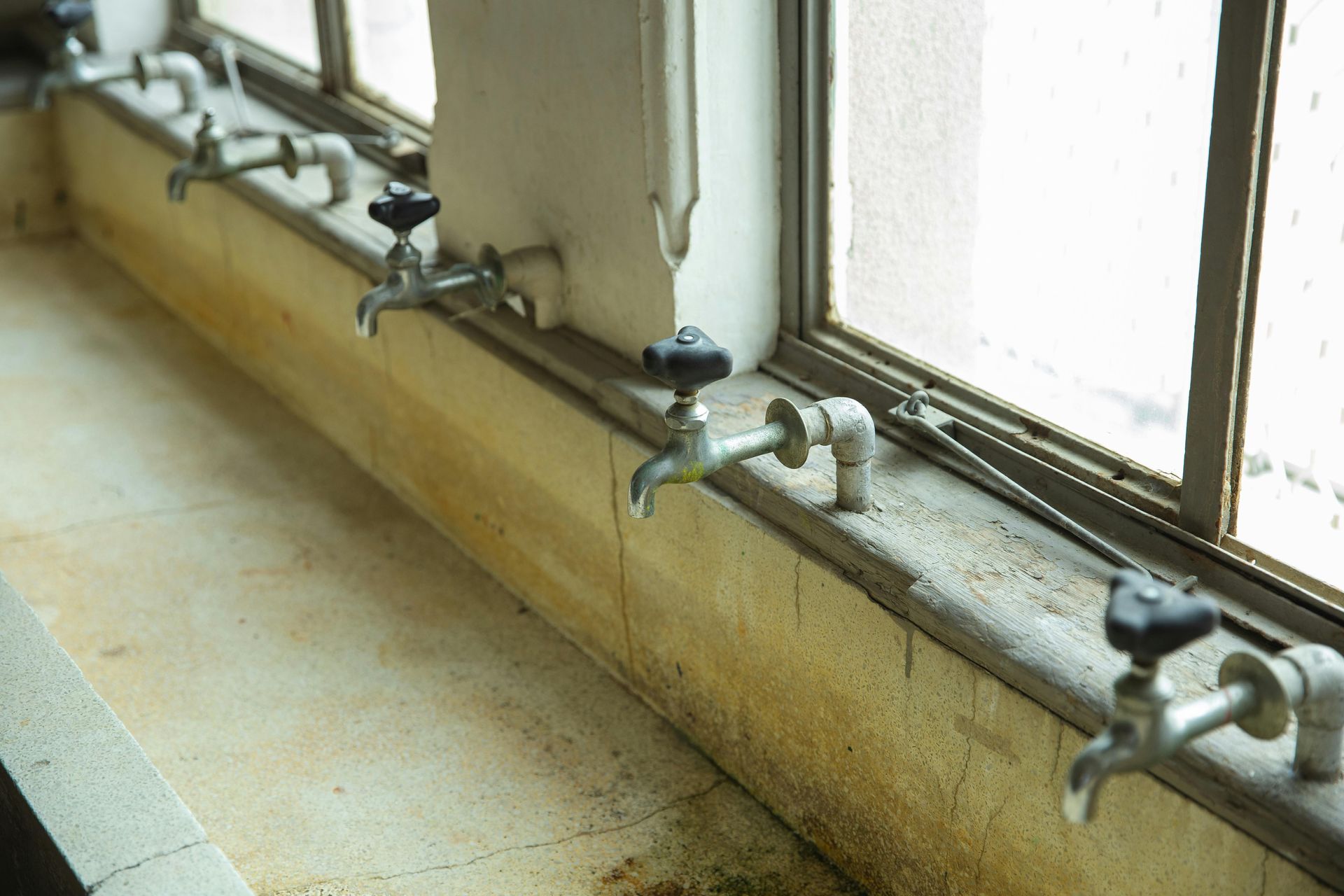 Row of water taps along a windowsill in a room with peeling paint and a window.