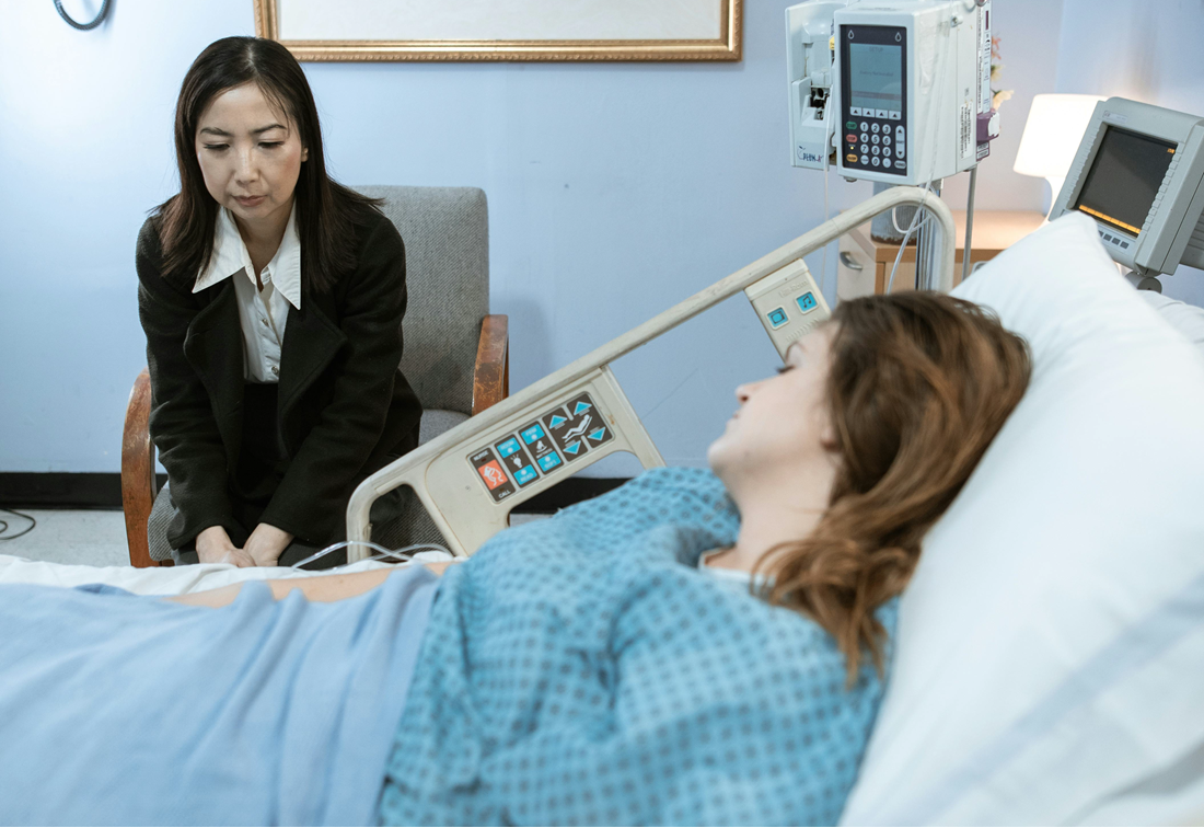 Visitor beside a hospital bed, speaking with a resting patient in a blue gown