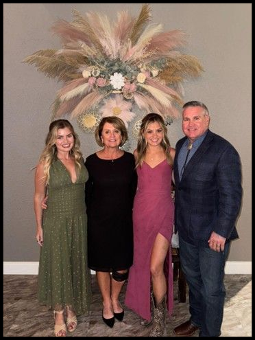 Four people stand side-by-side posing for a photo in front of a decorative pampas grass display against a gray wall.