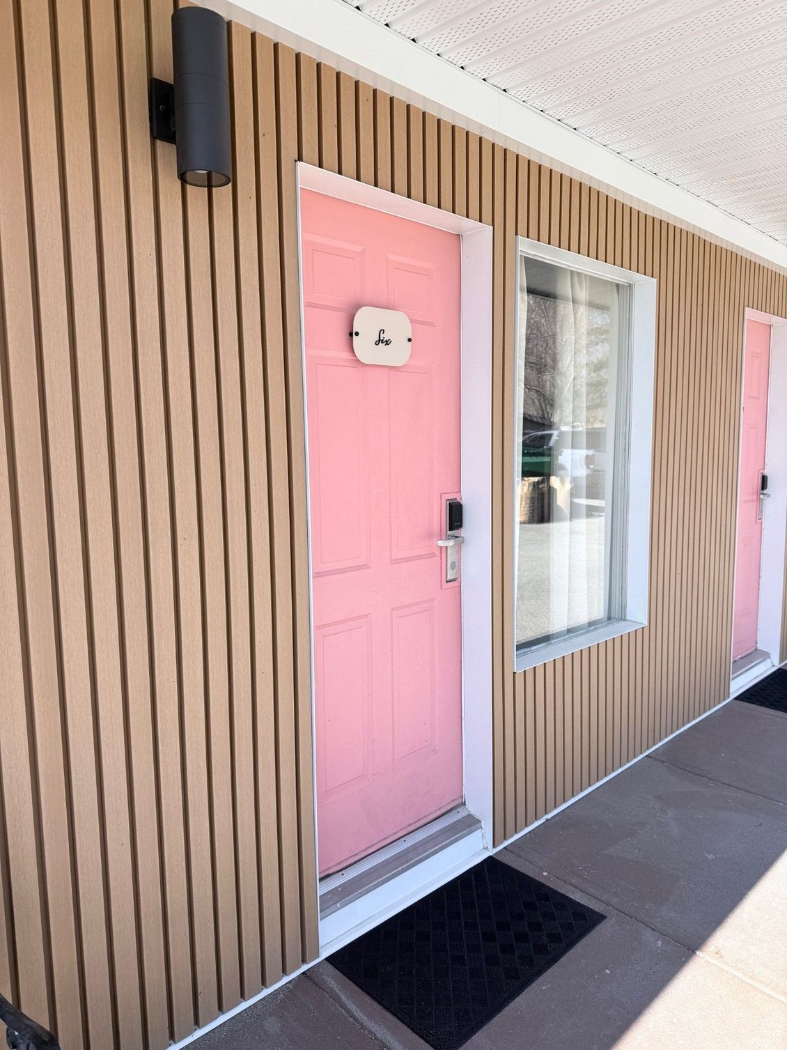 A bright pink door with a digital lock and a wall-mounted light on a building exterior with tan vertical wood siding.