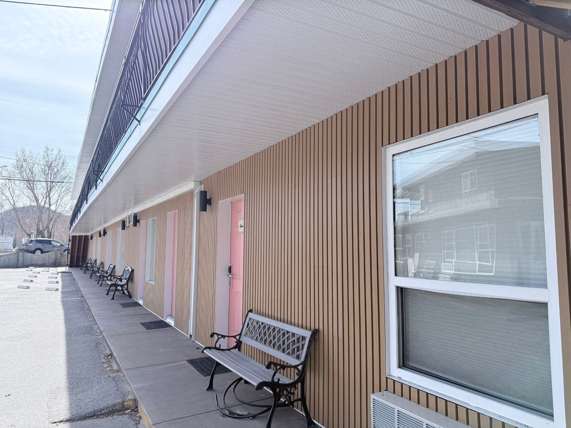 A low-angle view of a motel exterior with vertical tan siding, a long walkway, and outdoor benches under a porch overhang.