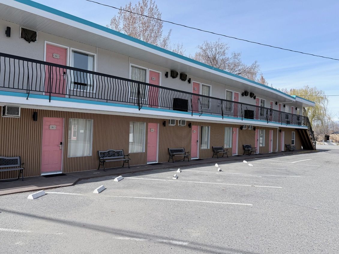Two-story motel with pink doors, light gray siding, blue trim, and a black metal railing along the upper walkway.