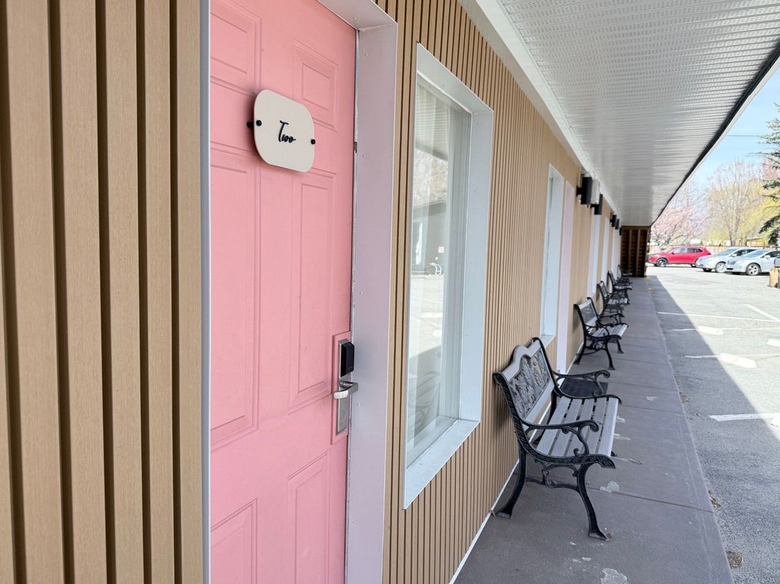 A row of motel rooms with pink doors and metal benches sits along a concrete walkway under a covered outdoor corridor.