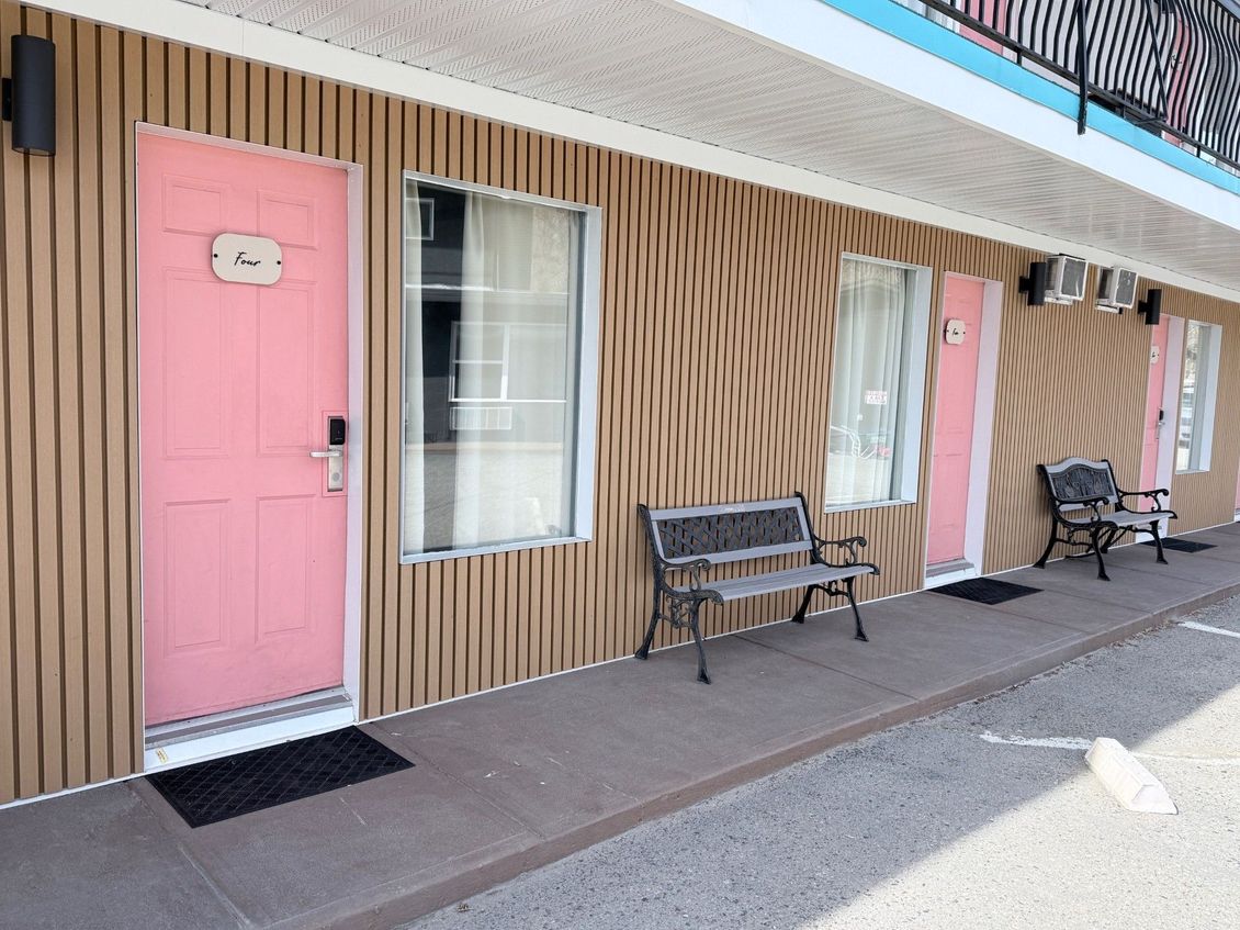 A row of motel units with pink doors, vertical tan wood siding, and metal benches on a concrete walkway.