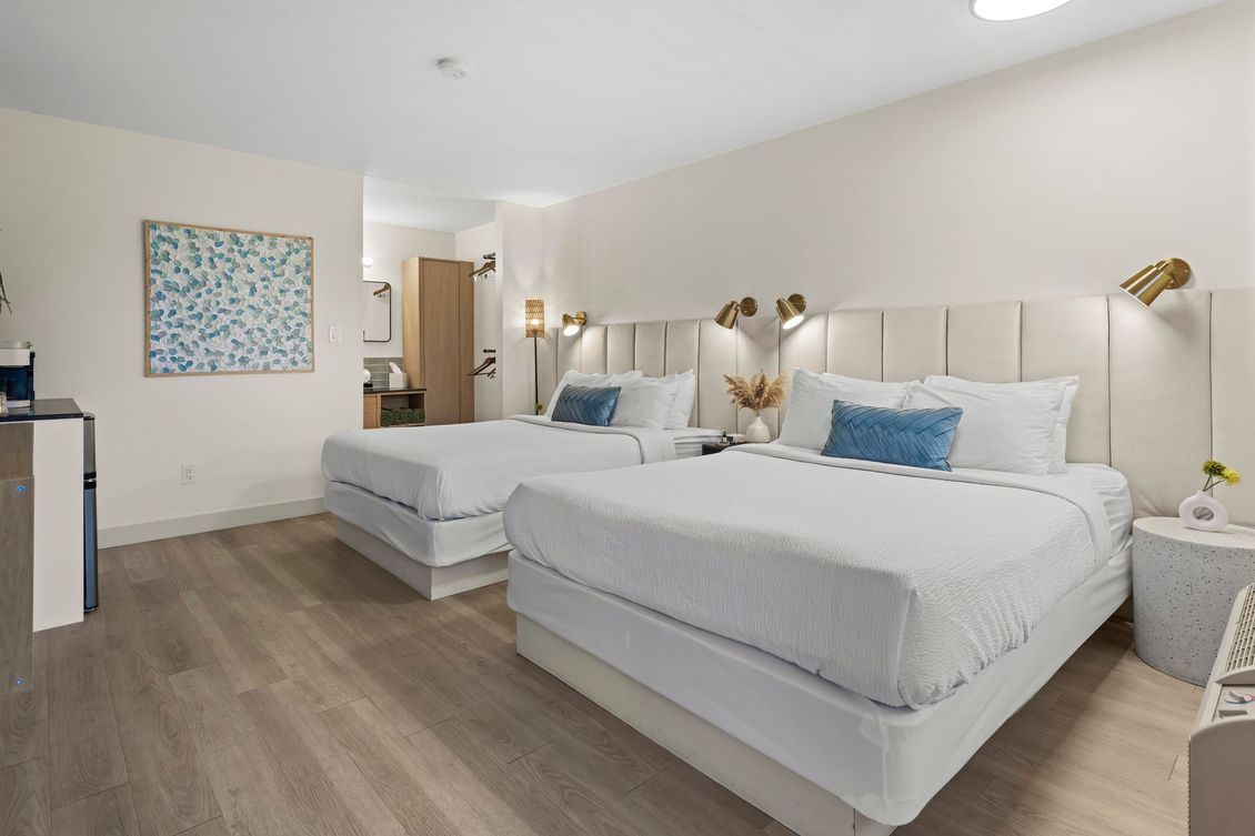 A modern hotel room featuring two queen-sized beds with white bedding, blue accent pillows, and neutral decor.