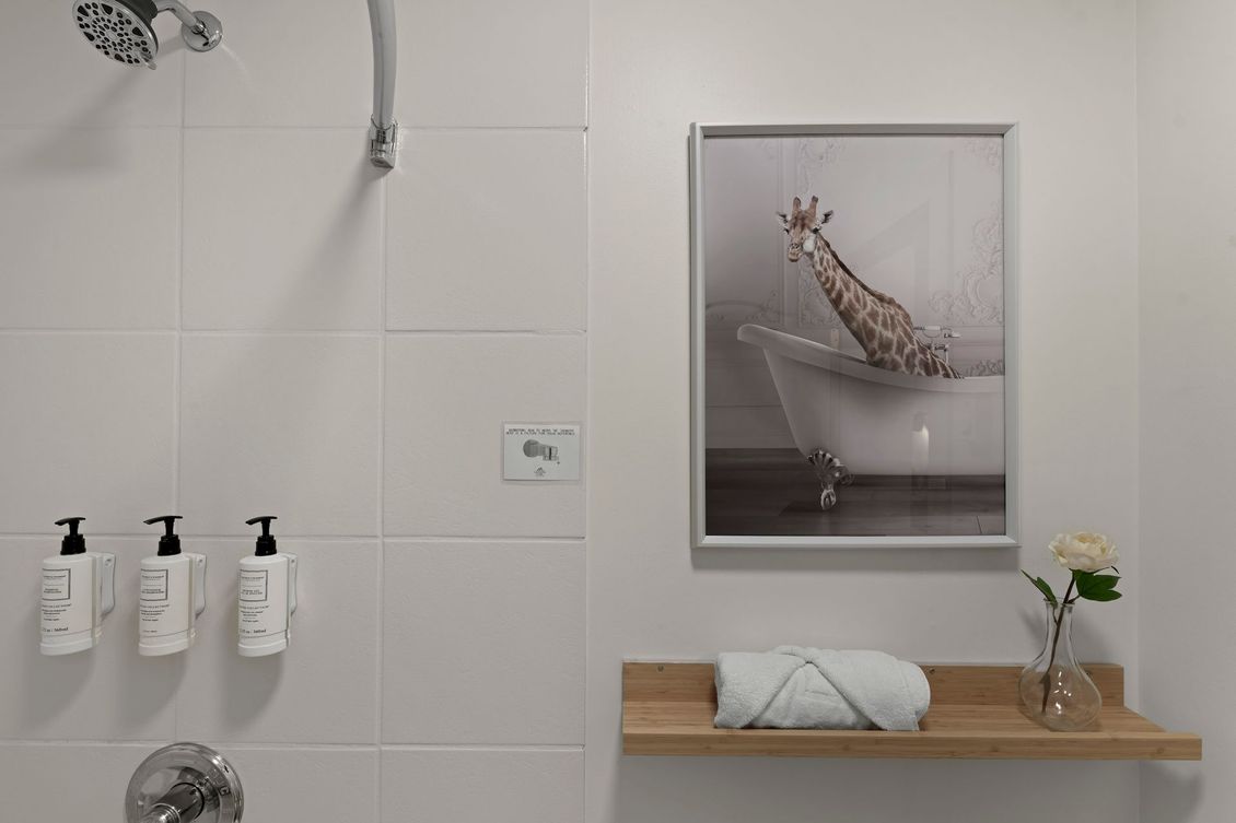 A white-tiled shower area with wall-mounted soap dispensers, a framed print of a giraffe in a tub, and a towel on a shelf.