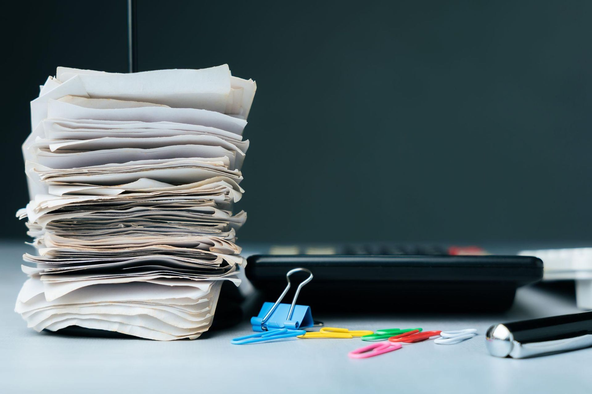 A stack of papers sits next to a calculator, a blue binder clip, colorful paper clips, and a pen on a desk.