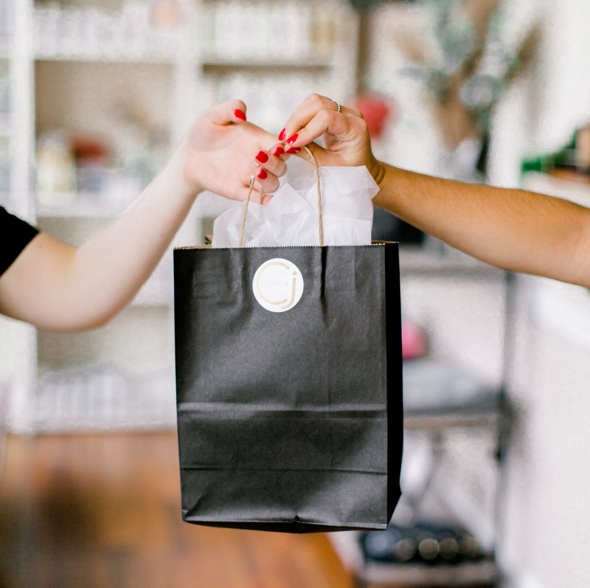 Two people exchanging a black paper shopping bag. One hand reaches out, the other holds the bag. White paper and logo visible.