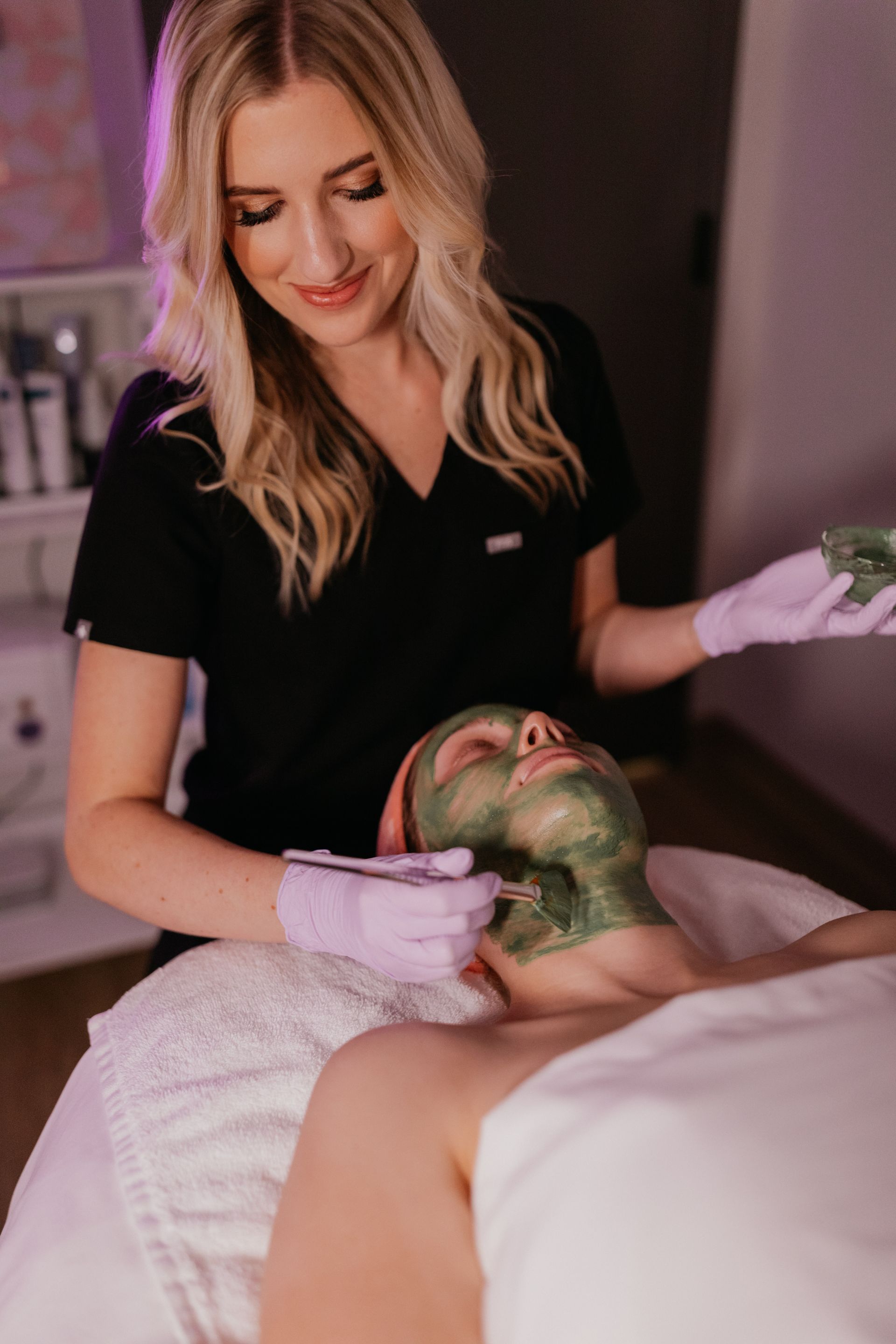 Aesthetician smiles, applying treatment to a client in a spa room. Blond hair, gloves, products on shelves.